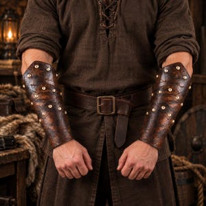 Puede incluir: Un par de brazaletes de cuero marrón oscuro con un acabado texturizado y desgastado, decorados con remaches metálicos en un patrón. Los brazaletes se llevan en los antebrazos, combinando con una túnica marrón oscuro y un cinturón de cuero.