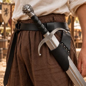May include: A silver-toned sword with a lion-head pommel and a black leather-wrapped grip, secured in a black leather scabbard attached to a wide black leather belt worn over brown trousers. The sword features a curved crossguard and a metal blade.