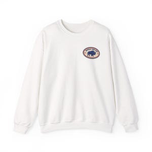 May include: A white crewneck sweatshirt featuring a small oval graphic on the left chest. The graphic contains a dark blue silhouette of a buffalo, the text "BUFFALO" and "NEW YORK" in dark blue, and a thin red border around the oval shape.