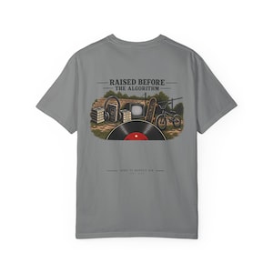 May include: A grey t-shirt featuring a graphic on the back with the text RAISED BEFORE THE ALGORITHM. The illustration includes a stack of cassette tapes, headphones, a television, a skateboard, a bicycle, and a large vinyl record in the foreground.