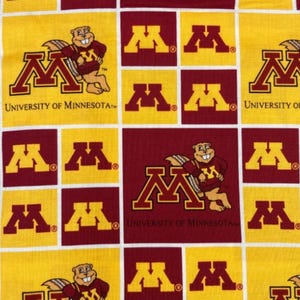 Könnte beinhalten: Ein Baumwollstoff mit einem wiederkehrenden Gittermuster aus kastanienbraunen und goldenen Quadraten. Das Design zeigt das Block-M-Logo der University of Minnesota und das Maskottchen Goldy Gopher. Ein Lineal links zeigt eine Skala von ca. 10 cm.