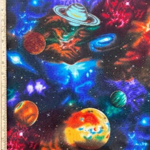 Könnte beinhalten: Ein Stoff mit einem lebendigen Weltraumdruck, der Planeten, Sterne und farbenfrohe Nebel in Blau-, Lila-, Orange- und Rottönen zeigt. Ein Holzlineal ist am linken Rand sichtbar und zeigt Messungen in Zentimetern bis zu 30 cm an.
