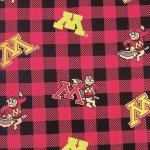 May include: Fabric featuring a red and black buffalo plaid pattern with repeating yellow block M logos and cartoon gopher mascots wearing red sweaters with a yellow M. A partial ruler is visible along the left edge showing centimetre markings.