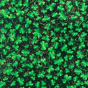 May include: A black fabric featuring a repeating pattern of vibrant green shamrocks and scattered gold glitter accents. A white ruler is visible along the left edge, showing markings in inches to provide a clear sense of scale for the printed design.