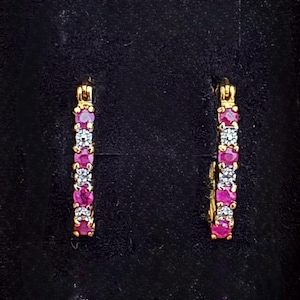 May include: A pair of gold hoop earrings featuring an alternating pattern of round-cut red rubies and clear white diamonds. The stones are set in a vertical row along the front of each hoop, creating a vibrant contrast between the gemstones.
