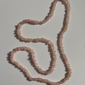 May include: A necklace composed of numerous small, irregular, pale pink rose quartz chip beads strung together. The beads have a translucent, crystalline appearance and are arranged in a continuous, flexible strand that forms a loose, organic shape.