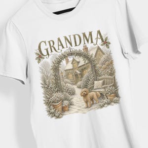 Pode incluir: Uma t-shirt branca com uma impressão gráfica de uma cena de jardim com neve, uma casa de campo, um banco e um cão de cor dourada. A palavra GRANDMA está impressa em letras grandes e decorativas douradas sobre a ilustração, acentuada com folhas de azevinho.