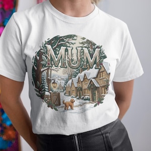 Könnte beinhalten: Ein weißes Kurzarm-T-Shirt mit einer kreisförmigen Grafik einer verschneiten Winterdorf-Szene mit Steinhäusern, einem goldbraunen Hund und dem Wort MUM in großen, stilisierten Buchstaben, die wie Baumzweige aussehen.