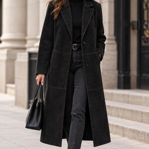 May include: A long black coat with a notched lapel and button closure, worn over a black turtleneck sweater and dark gray jeans with a black belt. The outfit includes black leather ankle boots and a black structured handbag with gold hardware.