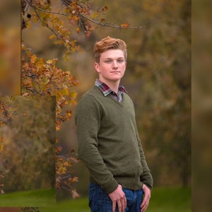 May include: A young man with red hair is wearing a green sweater and blue jeans. He is standing in front of a blurred background of fall foliage.