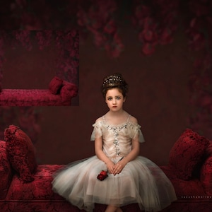Puede incluir: Una niña pequeña con un vestido blanco y una corona se sienta en un sofá de terciopelo rojo con una sola rosa roja en la mano. El sofá tiene dos almohadas y un fondo con estampado floral.