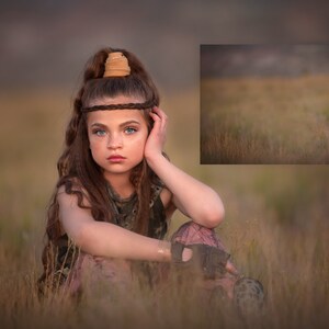 May include: A young girl with long brown hair sits in a field of tall grass. She is wearing a camouflage tank top and a brown leather headband. She has a serious expression on her face.