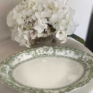 May include: An oval ceramic serving dish featuring a white centre and a wide, scalloped rim decorated with a detailed green floral pattern. The dish is positioned in front of a silver-toned metal bowl filled with a cluster of white hydrangea flowers.