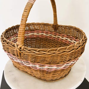 May include: An oval-shaped woven wicker basket featuring a single arched handle. The basket is constructed from natural brown reeds and includes decorative horizontal bands of light pink and white woven material around the upper body section.