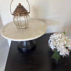 May include: A clear glass jar with vertical ribbing, enclosed in a brass-toned wire cage with a matching brass lid and a thin arched handle. The jar sits atop a white marble pedestal stand with a black base, next to a cluster of white flowers.