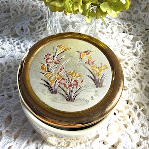 May include: A round ceramic jar with a gold-rimmed lid featuring a metallic silver inlay decorated with colorful irises and a bird. The side of the white base displays the text 24K GOLD TRIM in gold lettering, indicating the decorative finish.