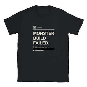 Puede incluir: Una camiseta negra de manga corta con texto en blanco que dice: BUILD REPORT, MONSTER BUILD FAILED. Missing body parts. RUNTIME BUREAU. El diseño está centrado en el pecho con líneas horizontales que enmarcan el texto y un icono.