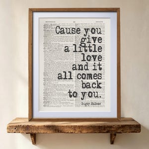 Può includere: Una stampa artistica incorniciata con uno sfondo di pagina di dizionario vintage e il testo nero sovrapposto: Cause you give a little love and it all comes back to you. Bugsy Malone. La stampa è inserita in una semplice cornice di legno con un passe-partout bianco.