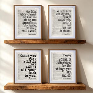 May include: Four framed prints featuring quotes from the movie Bugsy Malone printed on dictionary-style paper. The prints are displayed in wooden frames on two rustic wooden shelves. The text includes lines like Okay fellas, this is our moment, We could have been anything, Cause you give a little love, and You're gonna be remembered.