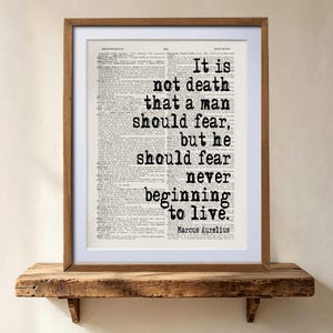 May include: A framed wall art print featuring a vintage dictionary page background with the black text: It is not death that a man should fear, but he should fear never beginning to live. Marcus Aurelius. The frame is made of medium-toned wood.