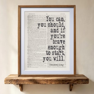 May include: A framed wall art print featuring a vintage dictionary page background with the black text: You can, you should, and if you're brave enough to start, you will. Stephen King. The print is enclosed in a simple wooden frame with a white mat.