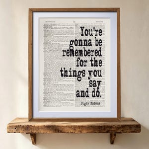 May include: A framed print featuring a vintage dictionary page background with the black text: You're gonna be remembered for the things you say and do. Bugsy Malone. The artwork is enclosed in a simple wooden frame with a white mat border.