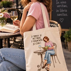 May include: A beige canvas tote bag featuring a printed illustration of a person sitting at a round table with a glass of wine and a book. The text printed on the bag reads: "У ВИНУ ЈЕ ИСТИНА, У КЊИГАМА ЈЕ СПАС!" The illustration shows a person wearing a pink shirt and blue jeans.