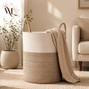 Peut inclure: Un grand panier de rangement cylindrique en corde tressée, présentant un design bicolore avec une partie supérieure crème et une partie inférieure beige. Le panier est doté de deux anses intégrées et d'une couverture en tricot beige drapée sur un côté.