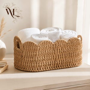 May include: An oval-shaped woven seagrass basket with a scalloped top edge and two integrated side handles. The basket is filled with several rolled white towels. The basket features a natural tan color and a textured, braided weave pattern.