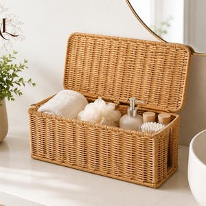 May include: A rectangular light brown woven storage basket with a hinged lid. The basket contains a white folded towel, a white bath sponge, a clear pump bottle with a silver nozzle, and two small containers holding white cotton swabs.