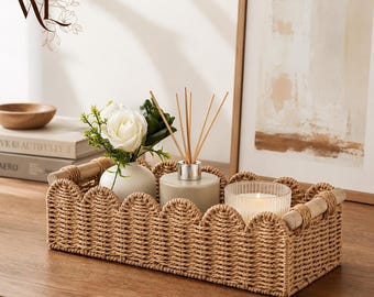 May include: A rectangular woven seagrass basket with a scalloped top edge and light wood handles. Inside the basket sits a small white vase with white artificial roses, a beige reed diffuser with wooden sticks, and a ribbed glass candle holder.