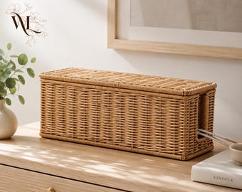 May include: A rectangular light brown woven wicker cable management box with a removable lid. The box features a horizontal weave pattern and a side opening for routing electrical cords. The structure is elongated and designed for storage.