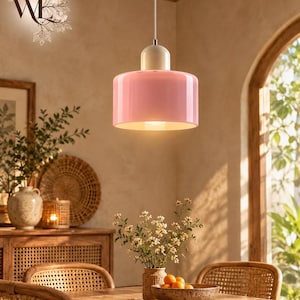 May include: A cylindrical pendant light fixture featuring a glossy pastel pink glass shade and a smooth cream-colored top cap. The lamp hangs from a thin silver cord, emitting a warm glow from the interior, creating a soft illumination.