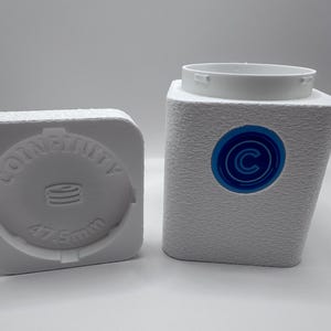 May include: A white, square-shaped storage container with a textured exterior and a circular opening at the top. A blue circular logo with a white letter C is on the front. Beside it, a matching white lid features the text COINFINITY and 47.5mm.