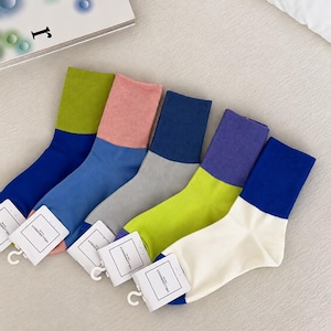 Pode incluir: Cinco pares de meias de cano médio com designs color-block. Cada par apresenta uma combinação bicolor distinta: verde-limão e azul, rosa seco e azul, azul-escuro e cinzento, roxo e verde-limão, e azul e branco-sujo. Cada par tem uma etiqueta.