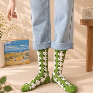 May include: A pair of mid-calf socks featuring a repeating pattern of white and green organic shapes outlined in black. The socks are worn with light blue denim jeans that are cuffed at the bottom, showcasing the full length of the patterned hosiery.