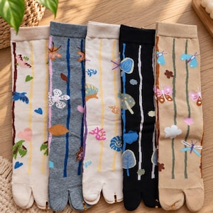 Puede incluir: Cinco pares de calcetines de dedo con rayas verticales y diversos motivos naturales, como mariposas, setas, flores y hojas. Los calcetines se presentan en colores crema, gris, beige, negro y marrón claro con diseños diferenciados.