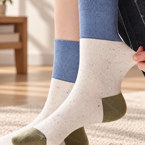 Può includere: Un paio di calze a metà polpaccio con base color crema e macchioline multicolori. Le calze presentano un bordo a coste blu a tinta unita nella parte superiore e una sezione del tallone e della punta in verde oliva, creando un design.