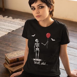 May include: A black short-sleeved t-shirt featuring a white graphic of a skeleton holding a red balloon with two small white bats flying above. The text "Well, shit..." is printed in white script font below the skeleton illustration.