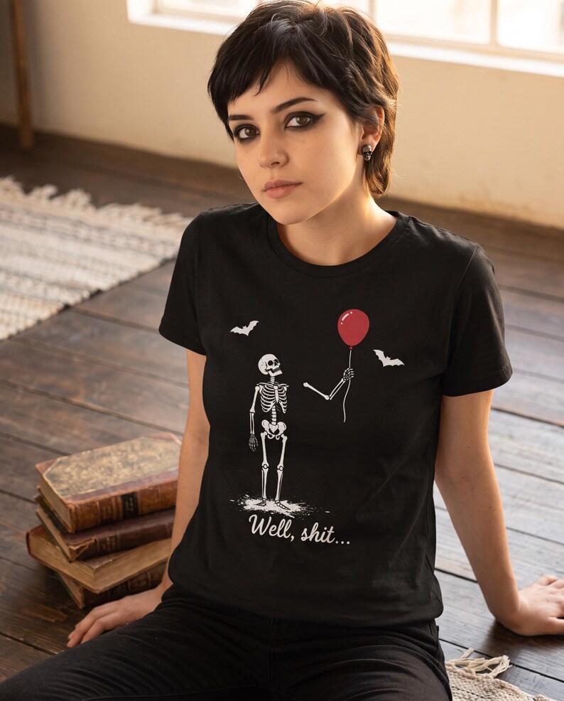May include: A black short-sleeved t-shirt featuring a white graphic of a skeleton holding a red balloon with two small white bats flying above. The text "Well, shit..." is printed in white script font below the skeleton illustration.