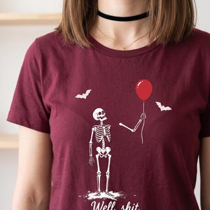May include: A maroon short-sleeved t-shirt featuring a white graphic of a skeleton holding a red balloon with two bats flying nearby. Below the skeleton, the text reads "Well, shit..." in a white cursive font, centred on the chest area.