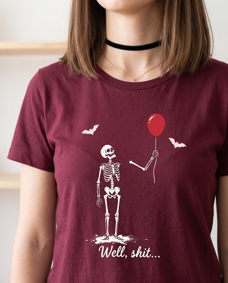 May include: A maroon short-sleeved t-shirt featuring a white graphic of a skeleton holding a red balloon with two bats flying nearby. Below the skeleton, the text reads "Well, shit..." in a white cursive font, centred on the chest area.