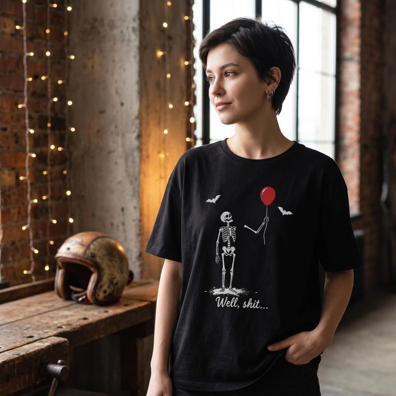 May include: A black t-shirt featuring a white graphic of a skeleton holding a single red balloon. The design includes two small white bats and the text "Well, shit..." printed in a white script font below the skeleton figure on the front of the shirt.