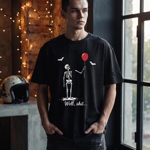 May include: A black short-sleeved t-shirt featuring a white graphic of a skeleton holding a single red balloon. Above the skeleton are two small white bats. Below the skeleton, the text "Well, shit..." is printed in a white cursive font.