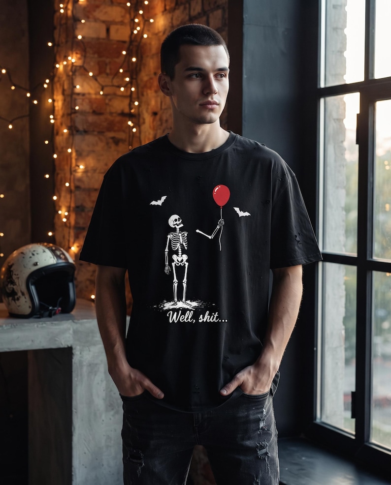 May include: A black short-sleeved t-shirt featuring a white graphic of a skeleton holding a single red balloon. Above the skeleton are two small white bats. Below the skeleton, the text "Well, shit..." is printed in a white cursive font.