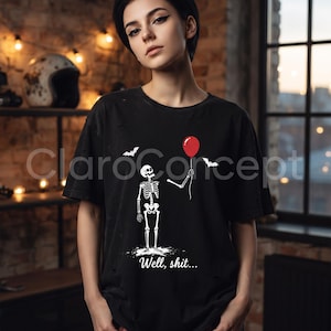 May include: A black t-shirt featuring a graphic of a white skeleton holding a single red balloon. The design includes two small white bats and the text "Well, shit..." printed in white below the skeleton. The fabric has a distressed, worn look.