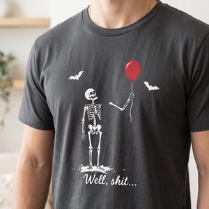 May include: A charcoal grey t-shirt featuring a white graphic of a skeleton standing and holding a single red balloon. Two white bats are flying in the background. The text "Well, shit..." is printed in white script font below the skeleton.
