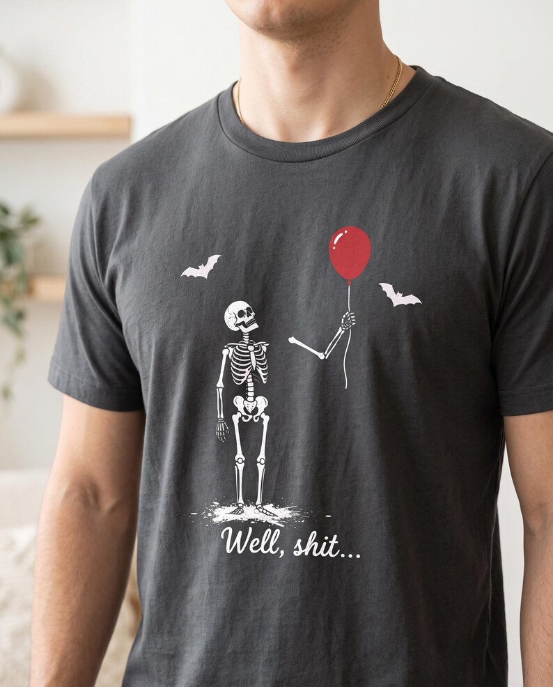 May include: A charcoal grey t-shirt featuring a white graphic of a skeleton standing and holding a single red balloon. Two white bats are flying in the background. The text "Well, shit..." is printed in white script font below the skeleton.
