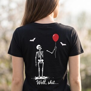 May include: A black short-sleeved t-shirt featuring a white graphic of a skeleton holding a red balloon. Two white bats are depicted above the skeleton. The text "Well, shit..." is printed in white script lettering at the bottom of the graphic.