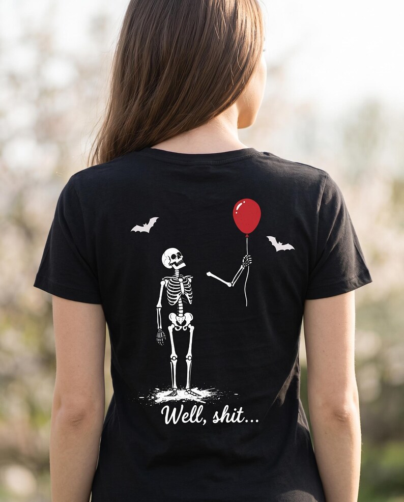 May include: A black short-sleeved t-shirt featuring a white graphic of a skeleton holding a red balloon. Two white bats are depicted above the skeleton. The text "Well, shit..." is printed in white script lettering at the bottom of the graphic.